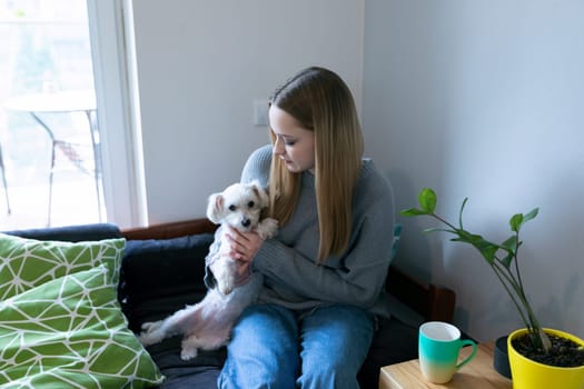 A young woman sits on a couch holding a small dog, enjoying a peaceful moment together in a bright, modern living room. Nearby, a coffee cup and plants add to the cozy atmosphere.