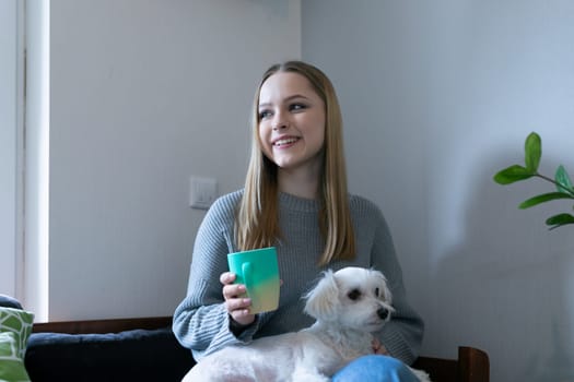 A young woman with long hair sits comfortably on a couch, holding a colored drink. Her small white dog snuggles beside her in a bright and inviting indoor space.