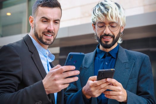 Two young businessmen in suits use their smartphones while standing outside in the city. They appear focused and engaged, reflecting modern communication and professional digital lifestyle.