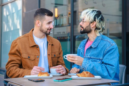 Two friends share a relaxed moment at an outdoor cafe, smiling and talking while drinking coffee and eating croissants. Casual setting with phones on table, reflecting modern urban lifestyle.
