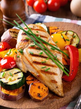 Freshly grilled chicken breast is placed atop a medley of colorful roasted vegetables including zucchini, bell peppers, and sweet potatoes. The dish is garnished with chives.