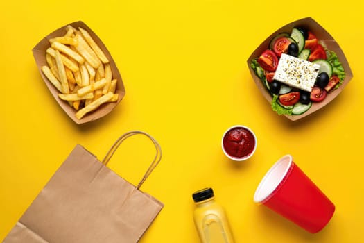 A colorful fast food spread features a box of golden fries, a fresh salad with tomatoes and olives, a red cup of drink, and a dipping sauce, all arranged on a bright yellow surface.