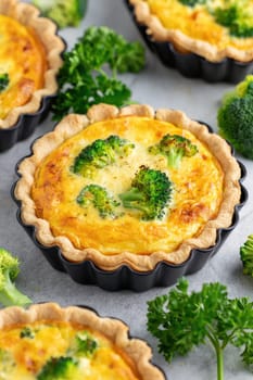 Broccoli tarts are freshly baked in round pans, showcasing a golden crust filled with creamy egg and vibrant green broccoli. Surrounding the tarts are fresh parsley and broccoli florets.