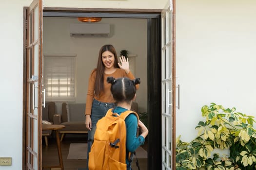 A cheerful mother stands at the door, waving her daughter off to school as she turns back to say goodbye. Their joyful exchange reflects the excitement of a new journey.