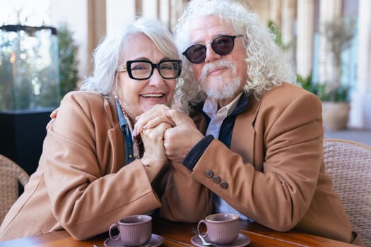Elderly couple with gray hair and glasses sitting at a cafe, sharing a smartphone while enjoying coffee.Dressed in matching brown coats, they smile, embracing technology in a stylish looking at camera