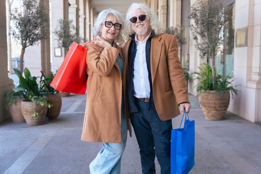 Elderly caucvasian couple smiling at the camera, hugging and holding vibrant shopping bags. Dressed in stylish coats, they enjoy a cheerful moment in an elegant outdoor shopping area. Vertical