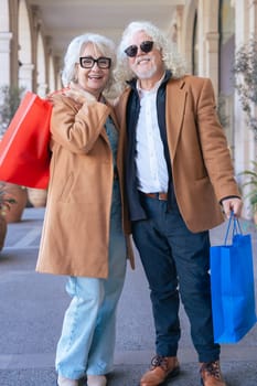 Elderly caucvasian couple smiling at the camera, hugging and holding vibrant shopping bags. Dressed in stylish coats, they enjoy a cheerful moment in an elegant outdoor shopping area. Vertical