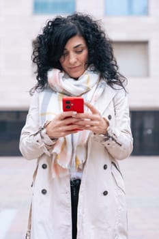 Happy caucasian woman standing outdoors in urban setting, wearing coat and scarf, using her red smartphone. She is smiling and relaxed, enjoying a casual moment on a cold day in the city. Vertical