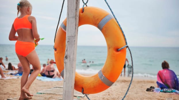 Orange lifebuoy hanging on wooden pole on a crowded beach with tourists enjoying summer vacation