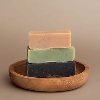 Three bars of natural handmade soap in soft, earthy colors are stacked neatly on a round wooden dish. The background is a plain, warm tone, enhancing the simple, elegant presentation.