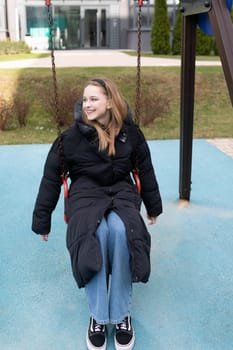 A young woman swings in a park, smiling joyfully. She wears a long black coat and jeans, enjoying a playful moment outdoors in a vibrant, green environment.