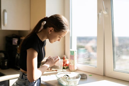 In a bright kitchen, a girl carefully adds spices to a bowl while focused on her cooking. Sunlight streams through the window, illuminating her concentration and ingredients.