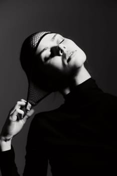 Graceful woman in a black turtleneck and fishnet headpiece, her eyes closed in serenity, soft lighting highlights her features against a dark background.
