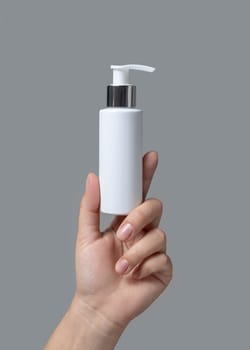 Female hand holding white cosmetic pump bottle against grey close up, package mockup, copy space. Skincare beauty product - lotion or cream, natural manicure