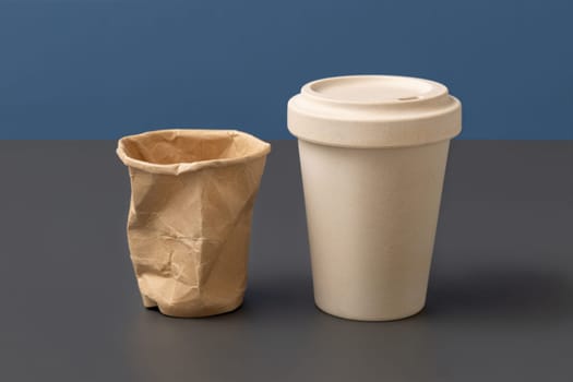 Used and crumpled disposable paper cup and reusable bamboo coffee cup on gray against blue closeup, copy space , mockup. Consumerism concept. No plastic and sustainable lifestyle concept