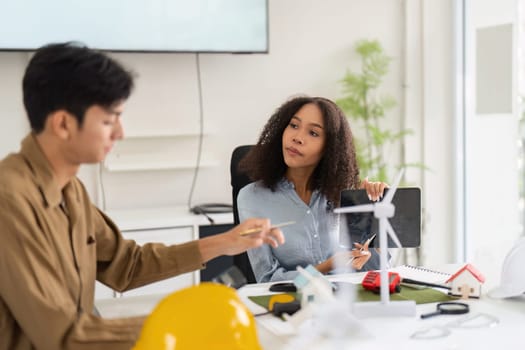 In a bright workspace, two professionals discuss projects involving renewable energy, showcasing models of wind turbines and energy-efficient homes.