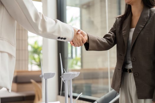 Amidst discussions of sustainability, two professional women share a handshake, featuring small wind turbines as a focal point of their agreement.