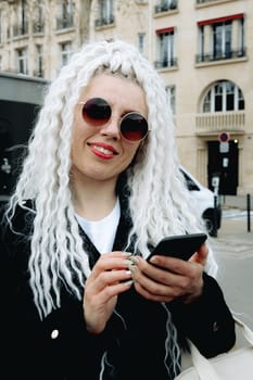 Portrait of one young beautiful Caucasian girl with a smile on her face in sunglasses with white afro curls holding a mobile phone in her hands and looking at the camera, in the city on a spring day, side view close-up. Concept using technology, city life, travel time.