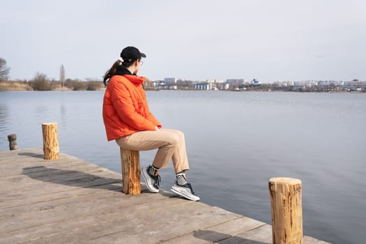 A young woman in an orange jacket sits on a wooden pier, gazing at a calm lake and distant cityscape on a sunny day.