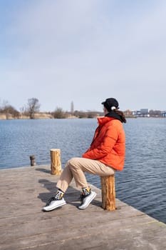 A young woman in an orange jacket sits on a wooden pier, gazing out at a tranquil lake on a sunny day.