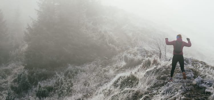 A woman athlete running along a rocky mountain trail in cold, misty weather, wearing a warm jacket, gloves, and a headband, showcasing endurance and determination.