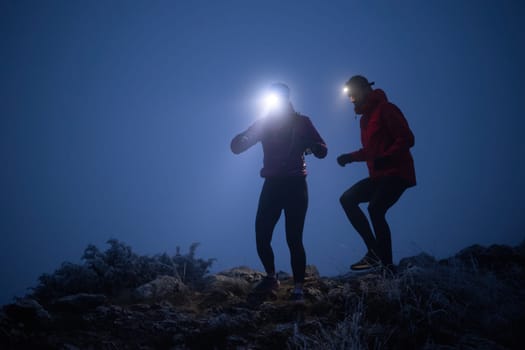 Two athletes run along a rugged mountain trail at night, wearing headlamps and winter gear, navigating the cold, misty terrain with determination and endurance.