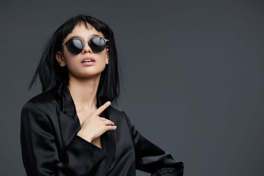 Woman, sunglasses, black blazer, gray background a woman in sunglasses and a black blazer poses confidently against a gray backdrop, hand on hip and finger on lips, exuding mystery and sophistication