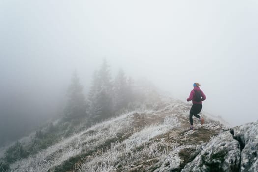 A woman athlete running along a rocky mountain trail in cold, misty weather, wearing a warm jacket, gloves, and a headband, showcasing endurance and determination.