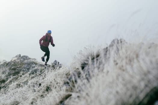 A woman athlete running along a rocky mountain trail in cold, misty weather, wearing a warm jacket, gloves, and a headband, showcasing endurance and determination.