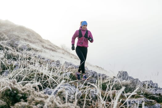 A woman athlete running along a rocky mountain trail in cold, misty weather, wearing a warm jacket, gloves, and a headband, showcasing endurance and determination.