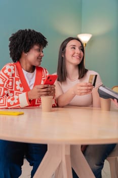 African American and Caucasian women smiling while paying at a coffee shop. One holds a card, the other a phone. Concept of friendship, technology, and casual lifestyle in a modern setting. Vertical