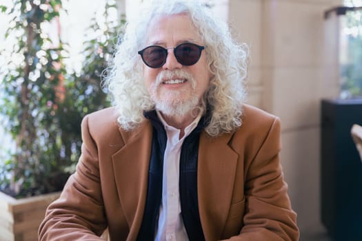 Confident senior man with white curly hair and sunglasses sitting at an outdoor cafe, smiling. Relaxed lifestyle, elegance, and sophistication in a modern setting with a cup of coffee and smartphone.