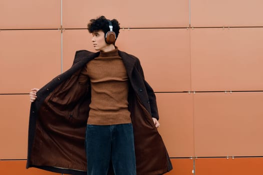 Fashionable young man in headphones and a coat standing against an orange wall. he is wearing a brown sweater, blue jeans, and brown shoes.