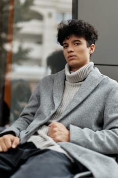 Fashionable young man in a grey coat and turtleneck sweater sitting on a bench in front of a window, focused on his phone.