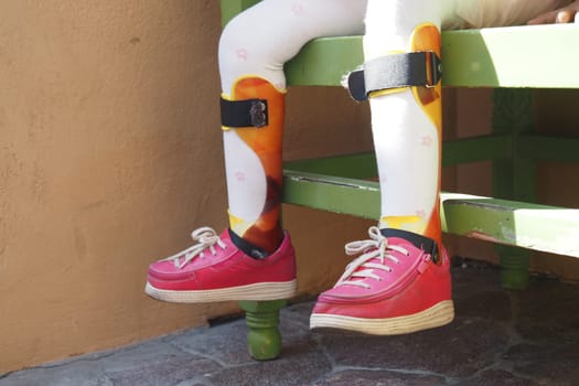 Child in pink shoes rests on a green bench with colorful leg braces.