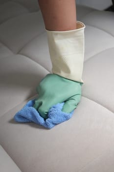 A hand wearing a glove wipes a sofa cushion with a cloth during cleaning.
