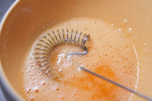 A silver whisk blends egg mixture with bubbles in a light bowl.