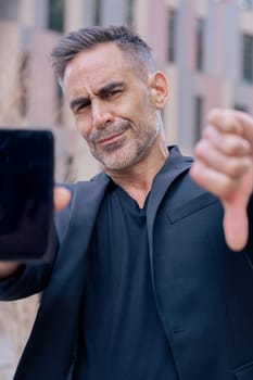Businessman in black outfit shows his smartphone and gives a thumbs down He serious confidently in an urban background, suggesting success and desapproval of modern technology looking at camera