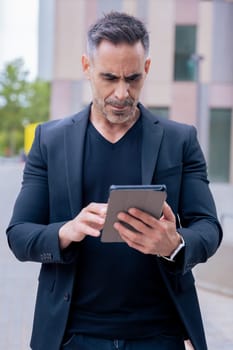 Confident businessman standing outdoors using a tablet in a modern urban setting.He appears focused and professional. Concept of digital work, technology, and contemporary business lifestyle. Vertical