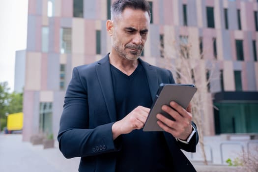 Confident businessman standing outdoors using a tablet in a modern urban setting. He appears focused and professional. Concept of digital work, technology, and contemporary business lifestyle.