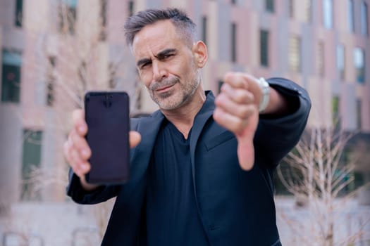 Businessman in black outfit shows his smartphone and gives a thumbs down He serious confidently in an urban background, suggesting success and desapproval of modern technology looking at camera
