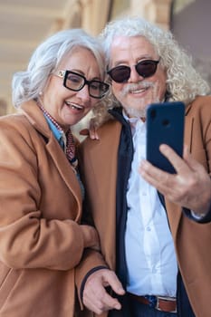 Caucasian senior couple smiling while using a smartphone together. They wear stylish jackets and glasses, enjoying a happy moment outdoors. Concept of love, connection, and modern lifestyle in age.