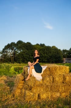 Young beautiful woman sits on a large pile of straw bales. Freedom, wind, setting sun