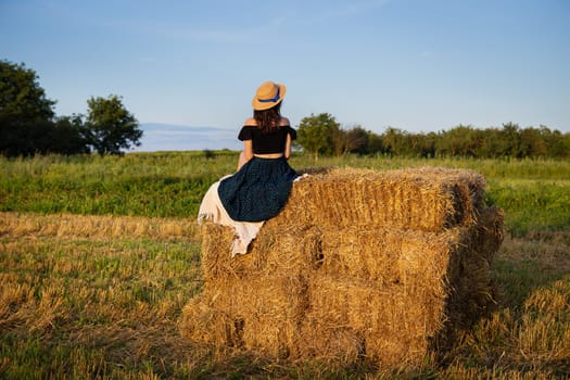 Young beautiful woman in a straw hat looks at the setting sun, the girl sits on a large pile of straw bales.