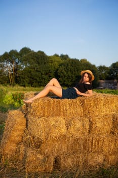A young beautiful woman in a straw hat lies on a large pile of straw bales and looks at the setting sun
