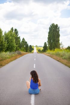 Attractive girl sitting on the asphalt and posing in the middle of the asphalt road near the field, copy space for content.