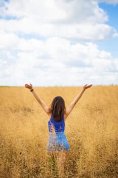Portrait of a young beautiful brunette woman in a blue T-shirt and denim shorts stands in the middle of the field, exposing her face to the sun on the background of a summer day outdoors