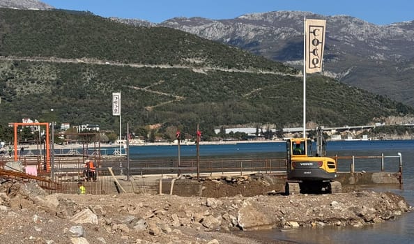 Budva,Montenegro-April 11,2025:A Volvo excavator working on a waterfront construction site with mountains and blue sea in the background. There is a flag with the word COCO next to it