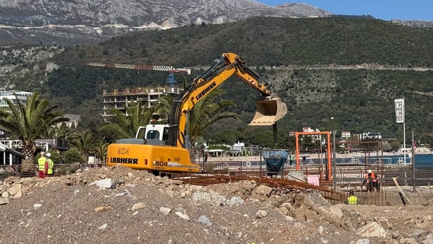 Budva,Montenegro-April 11,2025:Liebherr excavator operating at a beachside construction zone with visible rebar, workers, and palm trees. High quality photo