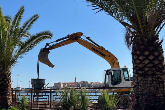 Budva,Montenegro-April 11,2025:A Liebherr excavator dumps material into a container at a coastal construction site lined with palm tree